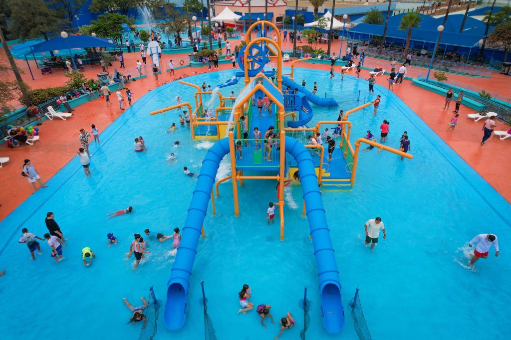 Vista aérea de alberca infantil con estructura de juegos acuáticos, toboganes azules y decenas de familias bañándose.