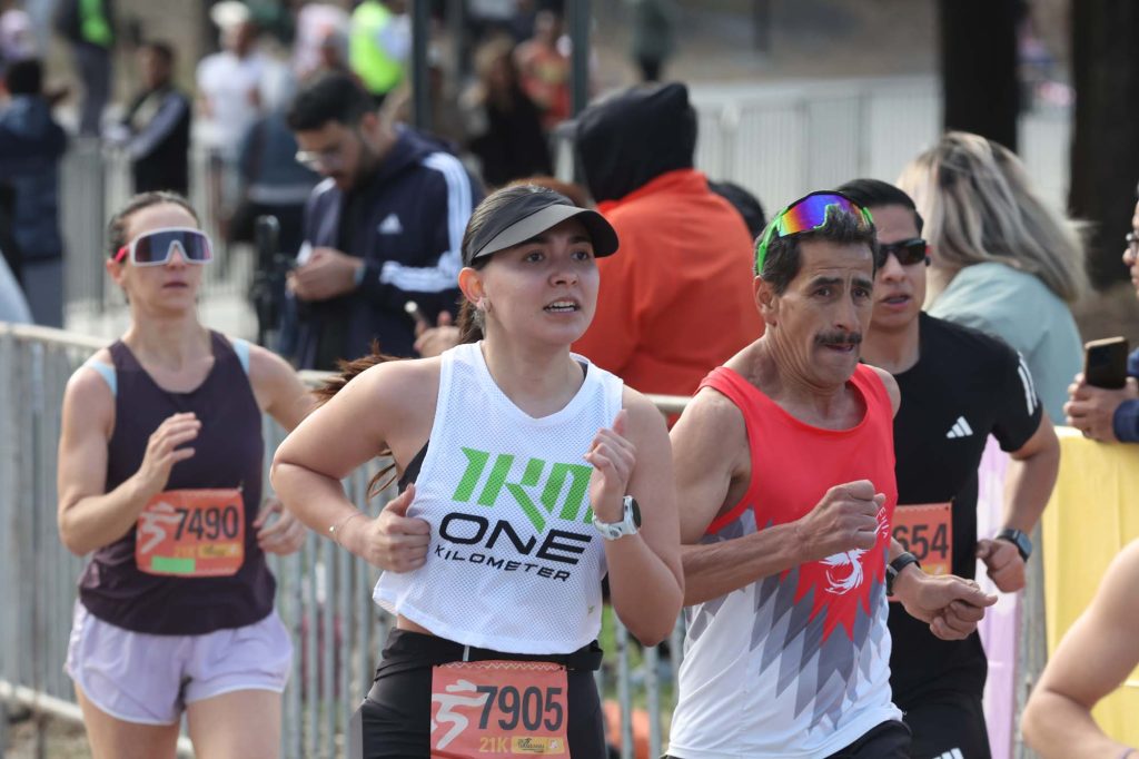 Corredores con números al pecho avanzan en una carrera de 21 kilómetros junto a vallas y público.