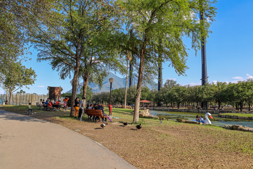 Familias descansan en el área verde junto a la laguna, con patos, lancha de cisne y chimeneas industriales.
