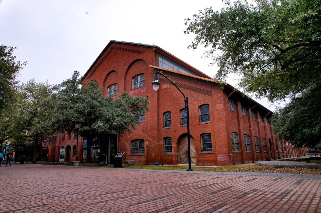 Nave industrial de ladrillo rojo rehabilitada, con ventanas de arco y explanada de adoquín entre árboles.