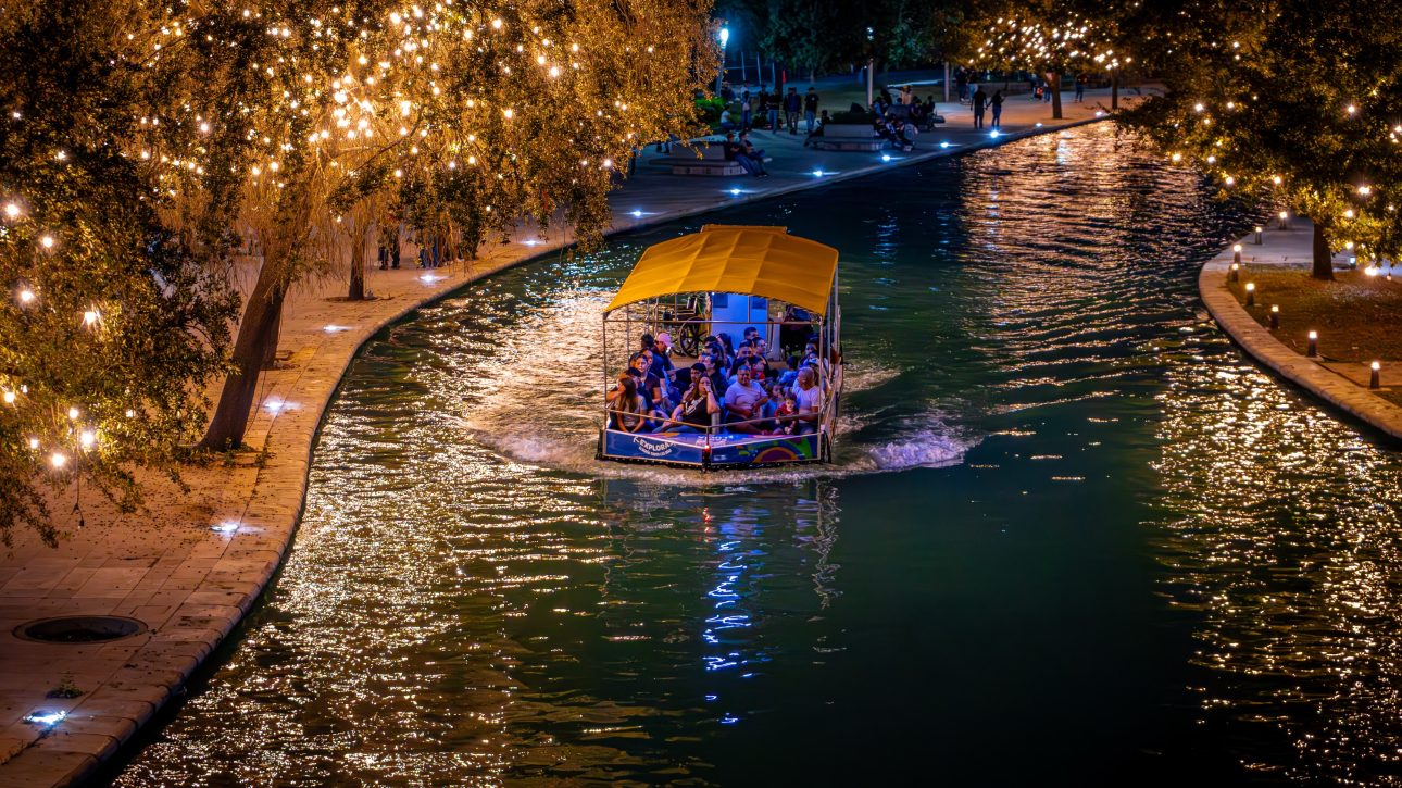 Barco turístico navegando de noche por el canal entre árboles cubiertos de luces cálidas