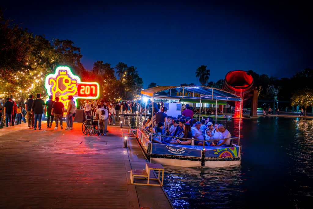 Embarcadero nocturno con visitantes, letrero de neón y barco turístico abordando en el canal